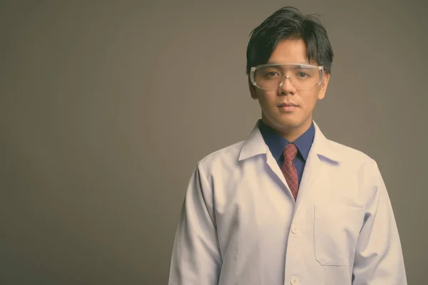 Молодий вродливий азіатський лікар з захисними окулярами на сірому фоні. — стокове фото