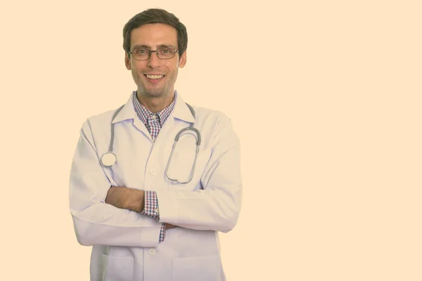 Studio shot of young happy man doctor smiling while wearing eyeglasses with arms crossed — Stock Photo, Image