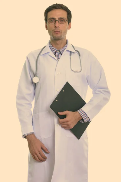 Studio shot of young handsome man doctor standing while holding clipboard — Stock Photo, Image