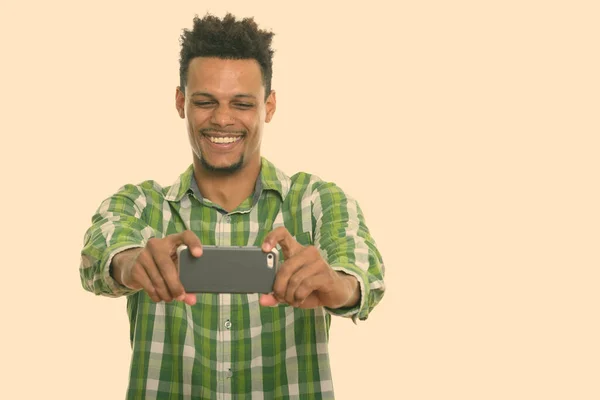 Estúdio tiro de jovem feliz homem africano sorrindo ao tirar foto com telefone celular — Fotografia de Stock
