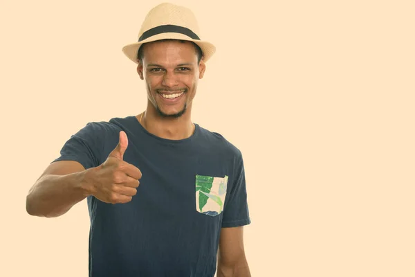 Estudio disparo de joven feliz africano sonriendo mientras llevaba sombrero y dando pulgar hacia arriba — Foto de Stock