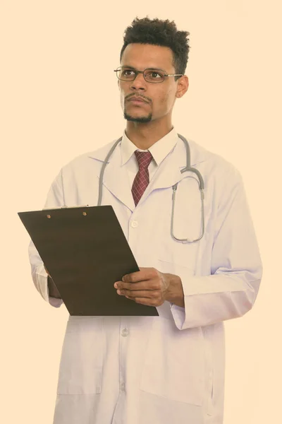 Studio shot of young African man doctor holding clipboard while thinking — Stock Photo, Image