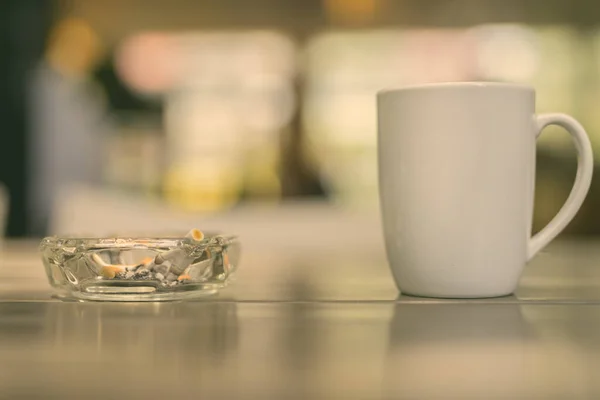 Coffee cup and ashtray with cigarettes on wooden table in restaurant with out of focus background and bokeh effect