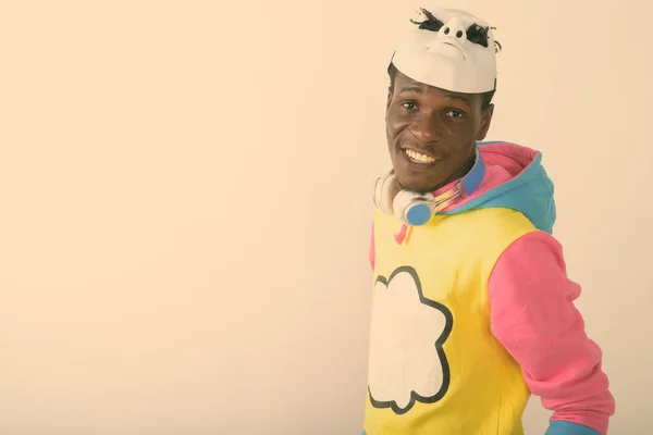 Estudio de disparo de joven feliz hombre africano negro sonriendo mientras llevaba máscara blanca en la parte superior de la cabeza y auriculares alrededor del cuello contra el fondo blanco — Foto de Stock