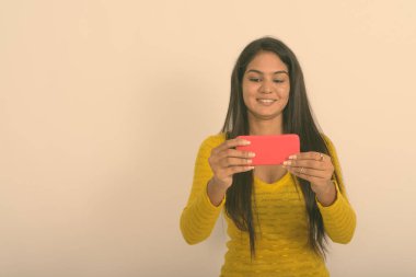 Cep telefonuyla beyaz arka planda fotoğraf çekilirken gülümseyen genç Hintli kadının stüdyo fotoğrafı.