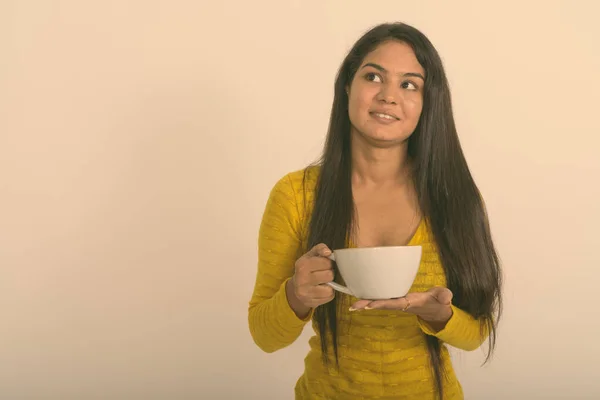 Estúdio tiro de jovem mulher indiana feliz sorrindo e pensando enquanto segurando xícara de café contra fundo branco — Fotografia de Stock
