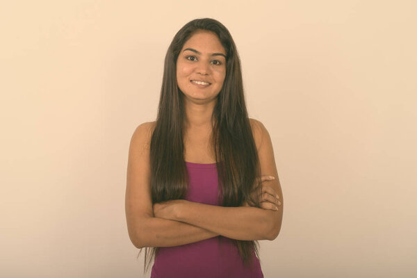 Studio shot of young happy Indian woman smiling while wearing sleeveless with arms crossed against white background