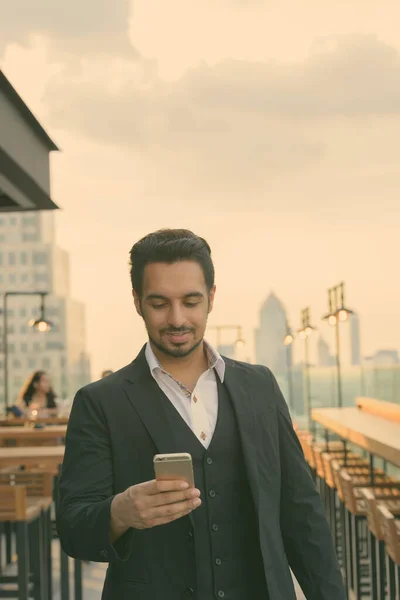 Jeune homme d'affaires indien heureux souriant tout en utilisant le téléphone mobile au restaurant sur le toit à Bangkok en Thaïlande — Photo
