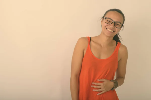 Estúdio tiro de jovem feliz mulher asiática sorrindo e rindo enquanto vestindo óculos e segurando o estômago contra fundo branco — Fotografia de Stock