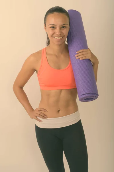 Genç, mutlu Asyalı bir kadının, gülümseyen ve ayakta duran yoga minderini beyaz arka planda spor için hazır tutarken çekilmiş bir fotoğraf. — Stok fotoğraf