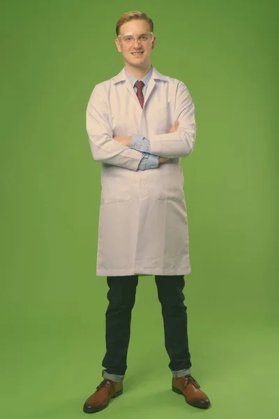 Joven hombre guapo médico con pelo rubio usando gafas protectoras contra fondo verde — Foto de Stock