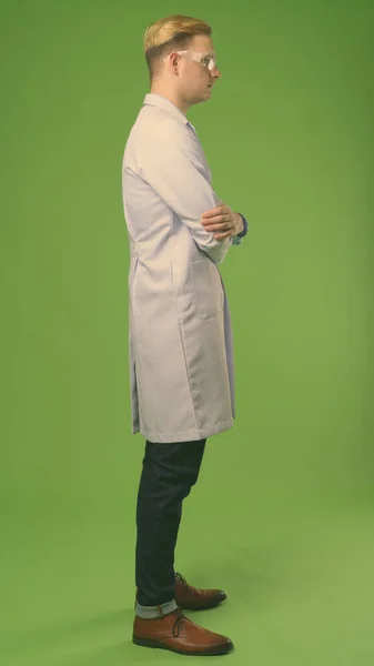 Νέος όμορφος άντρας γιατρός με ξανθά μαλλιά φοράει προστατευτικά γυαλιά σε πράσινο φόντο — Φωτογραφία Αρχείου