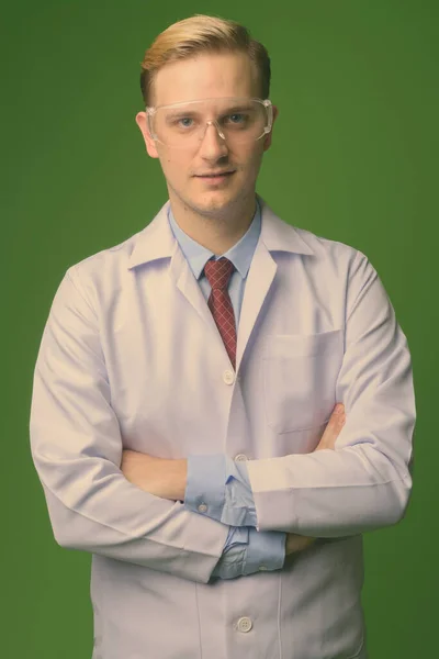 Mladý pohledný muž lékař s blond vlasy nosí ochranné brýle proti zelenému pozadí — Stock fotografie