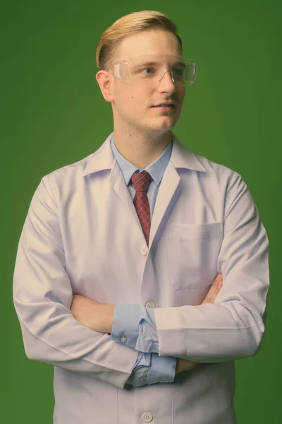 Joven hombre guapo médico con pelo rubio usando gafas protectoras contra fondo verde — Foto de Stock