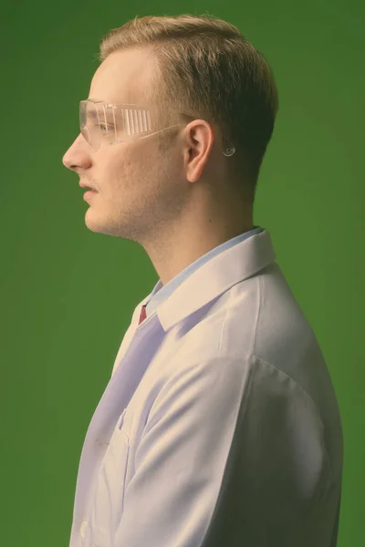 Joven hombre guapo médico con pelo rubio usando gafas protectoras contra fondo verde —  Fotos de Stock