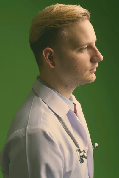 Young handsome man doctor with blond hair against green background — Stock Photo, Image