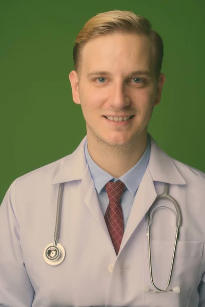 Young handsome man doctor with blond hair against green background — Stock Photo, Image