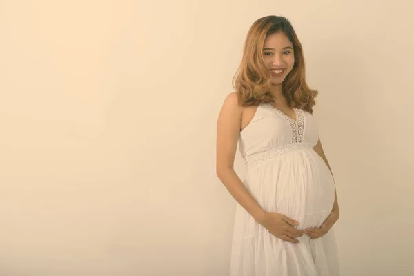 Studio shot młodej Azjatki w ciąży uśmiecha się trzymając brzuch na białym tle — Zdjęcie stockowe