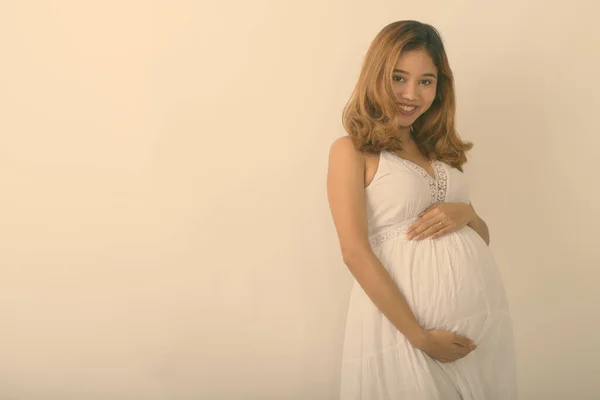 Studio shot of young happy Asian pregnant woman smiling while holding baby against white background — Stock Photo, Image