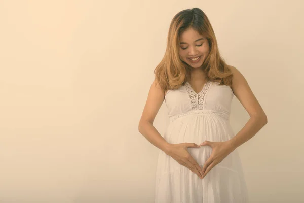Studio împușcat de tineri fericit asiatic femeie gravidă zâmbind în timp ce se uita la stomac și de a face semn de inima pe fundal alb — Fotografie, imagine de stoc