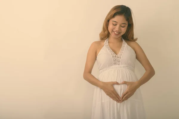 Studio shot de tineri fericit asiatic gravidă femeie zâmbind în timp ce face semn de inimă de mână pe stomac cu dragoste pentru copilul ei pe fundal alb — Fotografie, imagine de stoc