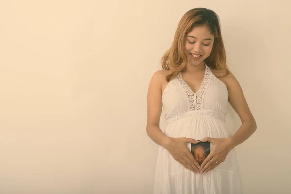 Studio împușcat de tânără femeie gravidă asiatică fericită zâmbind și uitându-se la sonograma copilului ei pe stomac pe fundal alb — Fotografie, imagine de stoc
