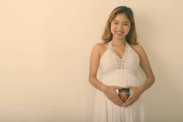 Studioaufnahme einer jungen glücklichen schwangeren Asiatin, die lächelt, während sie das Sonogramm ihres Babys auf dem Bauch hält und vor weißem Hintergrund ein Herzzeichen macht — Stockfoto