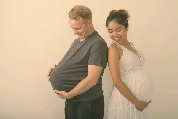 Studioaufnahme eines jungen glücklichen multiethnischen Ehepaares, das mit einem Mann lächelt, der Wassermelone als schwangeren Bauch vor weißem Hintergrund verwendet — Stockfoto