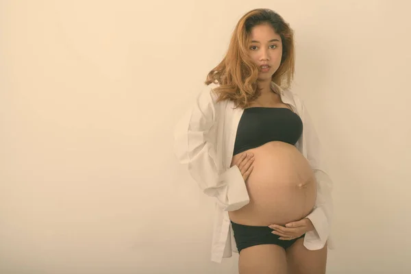 Studio împușcat de tânără femeie gravidă asiatică ținându-și copilul în timp ce purta cămașă albă deschisă pe fundal alb — Fotografie, imagine de stoc