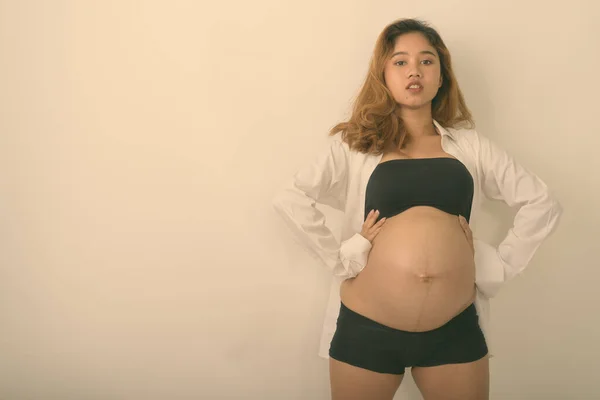 Studio împușcat de tânără femeie gravidă asiatică pozând în timp ce își ține copilul și poartă cămașă albă deschisă pe fundal alb — Fotografie, imagine de stoc