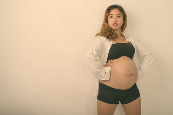 Genç Asyalı hamile kadının beyaz arka plana karşı bebeğini açık beyaz gömlekle tutarken düşündüğü ve poz verdiği bir fotoğraf. — Stok fotoğraf