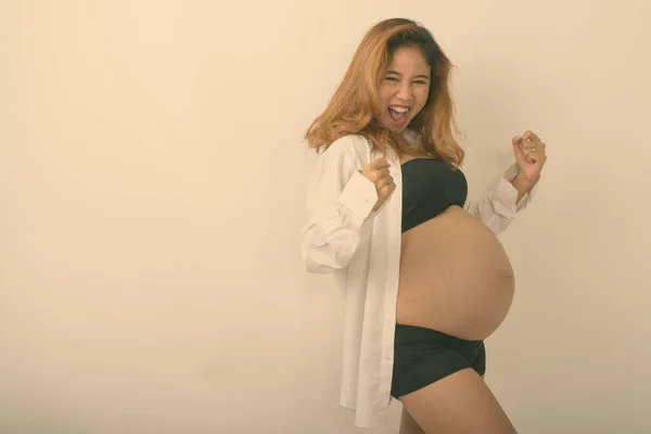 Estudio disparo de joven feliz mujer embarazada asiática sonriendo mientras que mira motivado y con camisa blanca abierta sobre fondo blanco —  Fotos de Stock