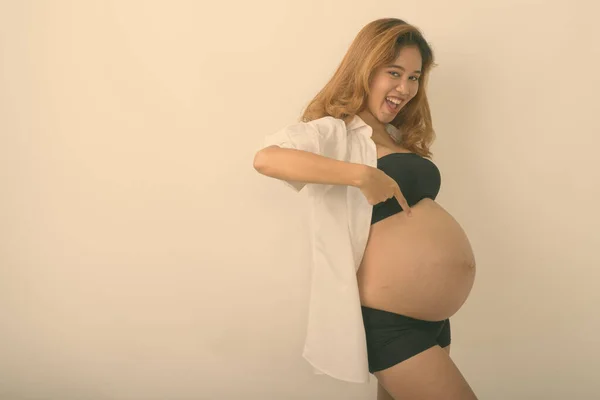 Studio shot de tineri fericit asiatic gravidă femeie zâmbind în timp ce arătând spre stomac și purtând cămașă albă deschisă pe fundal alb — Fotografie, imagine de stoc