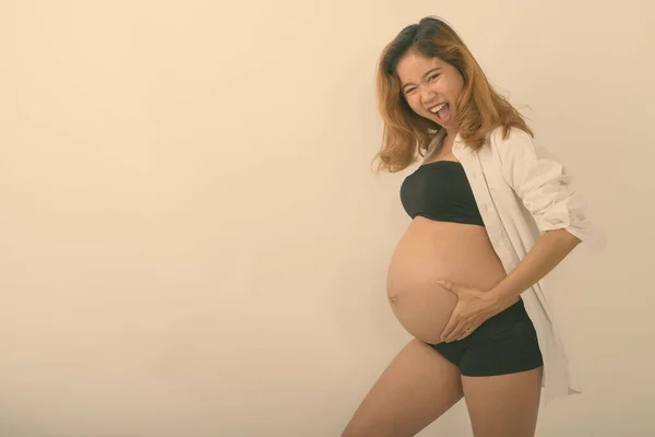 Studio shot de tineri fericit asiatic gravidă femeie zâmbind în timp ce ține stomacul și cu ochiul pe fundal alb — Fotografie, imagine de stoc