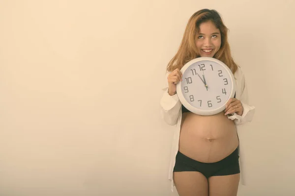 Studio shot de tineri fericit asiatic gravidă femeie zâmbind în timp ce ține ceas de perete cu ochii rostogolindu-se pe fundal alb — Fotografie, imagine de stoc