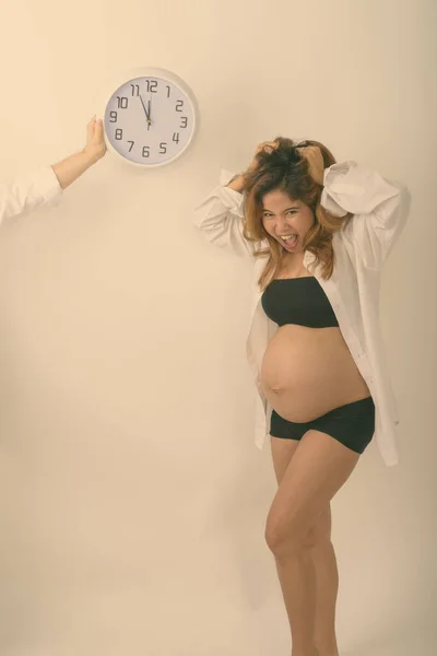 Студийный снимок молодой азиатской беременной женщины, выглядящей напряженной с обратным отсчетом на настенных часах на белом фоне — стоковое фото