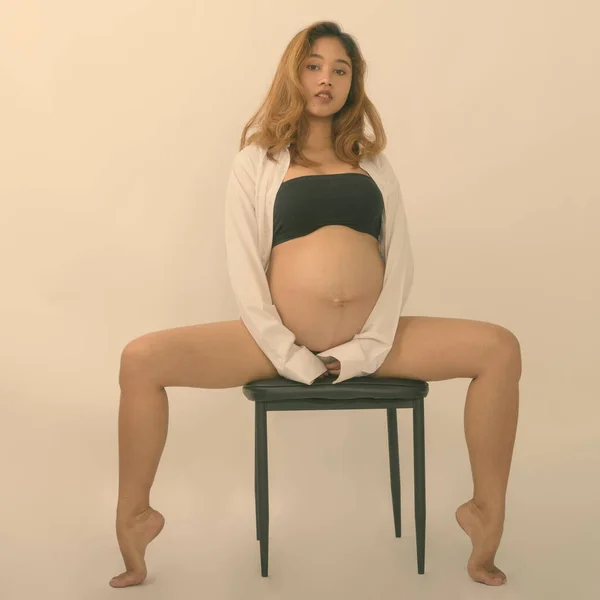 다리를 벌리고 하얀 배경을 배경으로 의자에 앉아 있는 젊은 아시아 임신부가 포즈를 취하는 모습의 전신 사진 — 스톡 사진