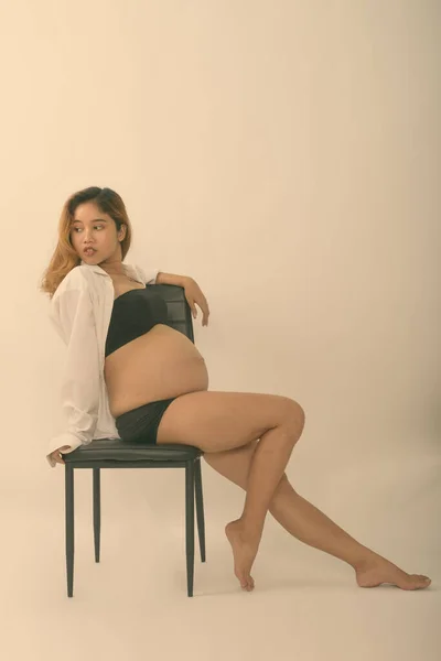 Ganzkörperaufnahme einer jungen schwangeren Asiatin, die auf einem Stuhl vor weißem Hintergrund sitzt und denkt und posiert — Stockfoto
