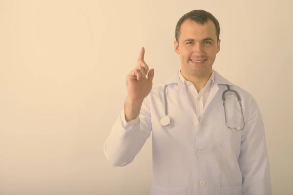 Estudio de tiro de joven feliz musculoso médico hombre sonriendo mientras señala el dedo hacia arriba contra fondo blanco — Foto de Stock