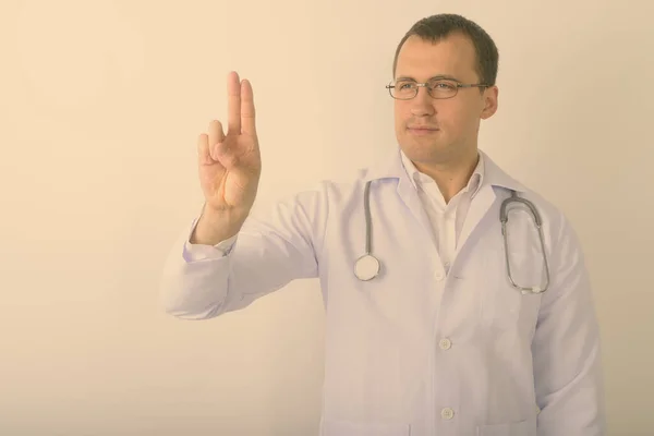 Estúdio tiro de jovem homem muscular médico pensando ao tocar em algo e vestindo óculos contra fundo branco — Fotografia de Stock