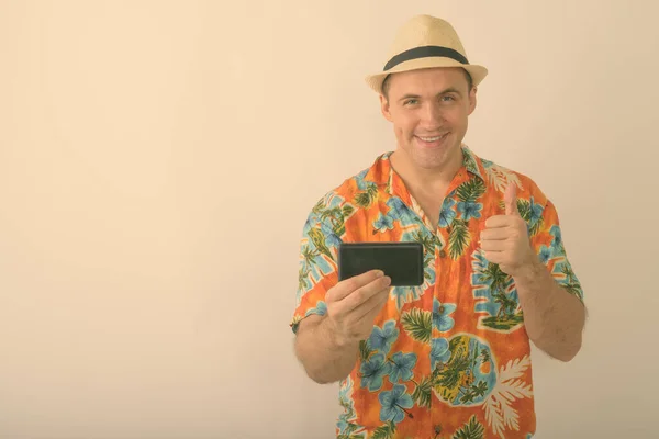 Студійний знімок молодого щасливого м'язового туриста, який посміхається, фотографувавши мобільний телефон і віддаючи великий палець на білому тлі — стокове фото