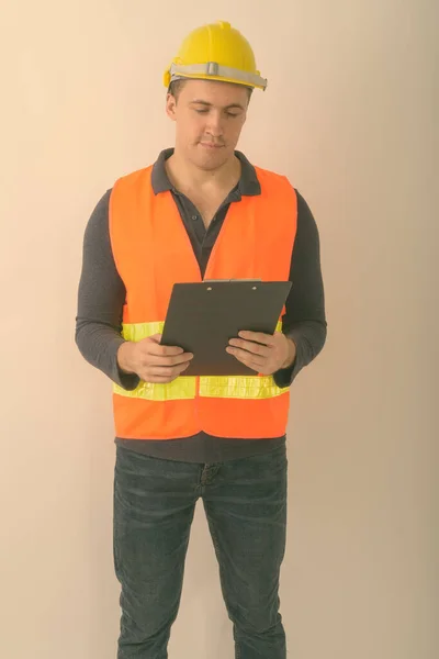 Studio shot of young muscular man construction worker standing and reading on clipboard against white background — Stock Photo, Image