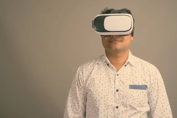 Young Indian businessman using virtual reality headset against gray background