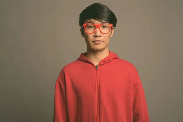 Jovem asiático vestindo casaco vermelho com óculos contra fundo cinza — Fotografia de Stock