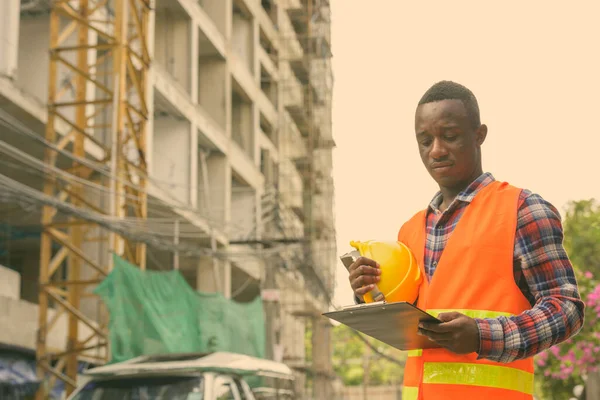 Jonge zwarte Afrikaanse man bouwvakker lezen op klembord terwijl het houden van harde hoed op de bouwplaats — Stockfoto