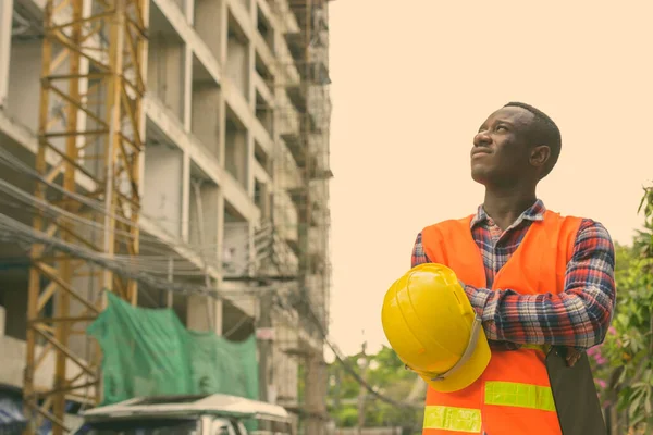 Pensativo joven africano negro trabajador de la construcción sosteniendo portapapeles y sombrero duro con los brazos cruzados en el sitio de construcción —  Fotos de Stock