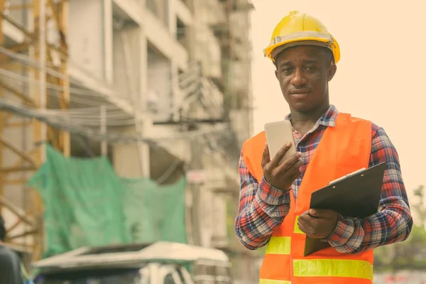 Jeune homme noir africain travailleur de la construction tenant presse-papiers et téléphone mobile sur le site de construction — Photo