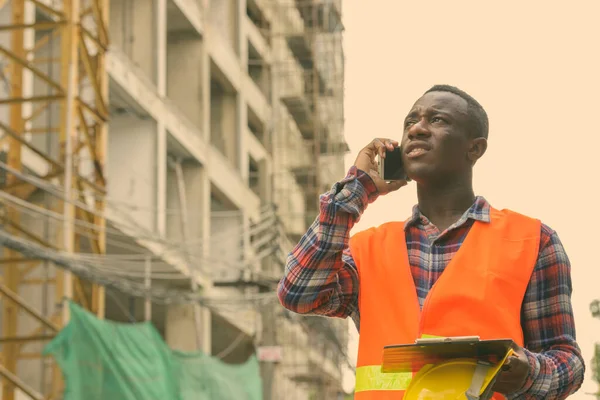 Bedachtzame jonge zwarte Afrikaanse man bouwvakker praten op mobiele telefoon terwijl het houden van veiligheidshelm en klembord op de bouwplaats — Stockfoto
