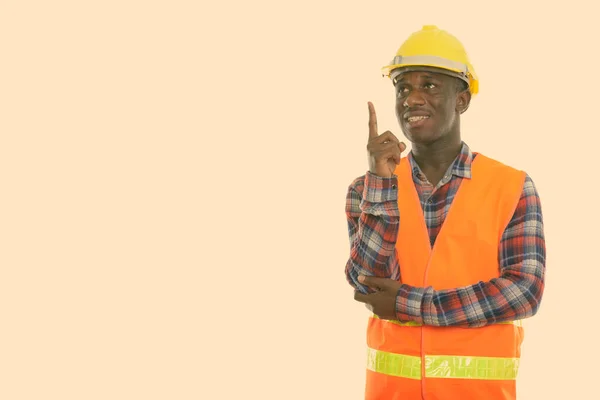 Studio shot van jonge gelukkige zwarte Afrikaanse man bouwvakker glimlachen en denken terwijl wijzend vinger omhoog — Stockfoto