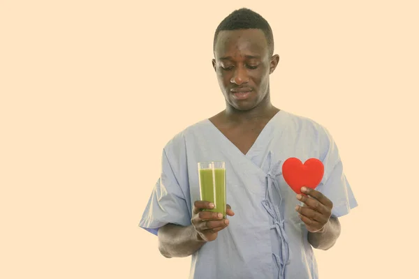 Estudio de un joven negro africano paciente mirando el vaso de batido de verduras verdes y sosteniendo el corazón rojo —  Fotos de Stock
